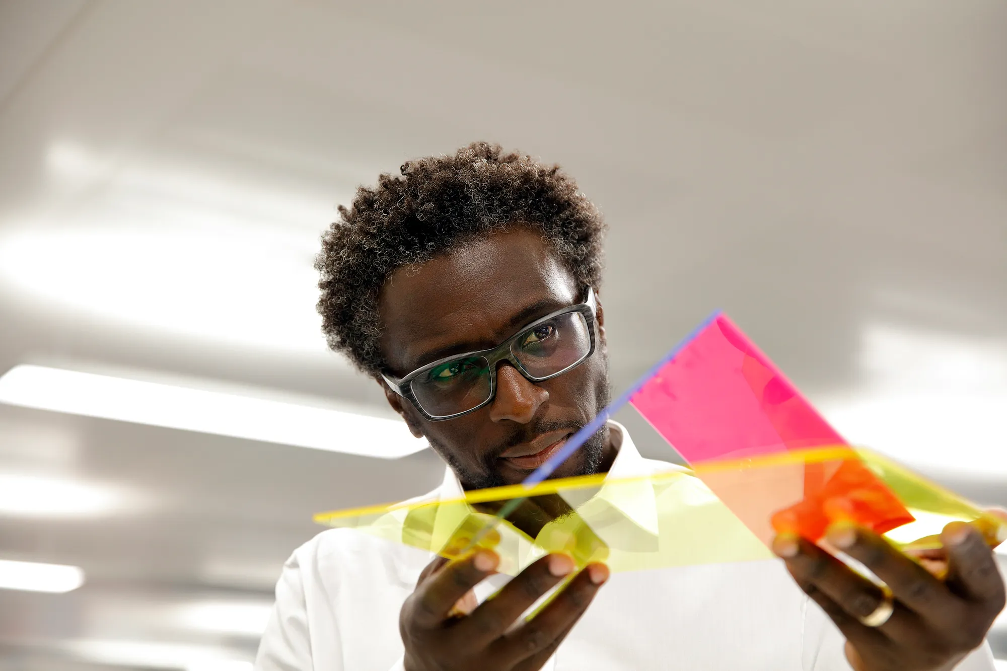 A man with glasses, wearing a white shirt, holds an array of colored plexiglass.